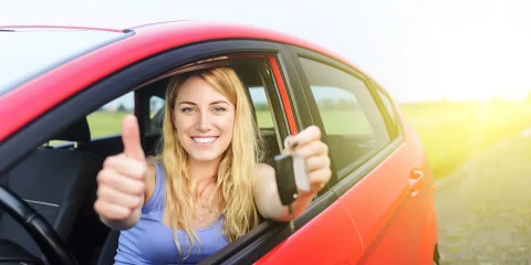 A woman inside a car holding keys with one hand and giving a thumbs up with the other hand, smiling.