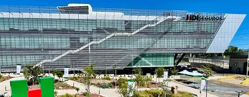 Exterior view of the HDI Seguros building on a sunny day, showing modern glass architecture and landscaped green areas in front. The webpage promotes HDI Seguros Mexico Tourist Auto Insurance.